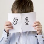 Ein Kind, das sein Gesicht hinter einem durchtrennten Papier versteckt. Auf einer Seite ist ein weibliches Strichmännchen gezeichnet auf der anderen Seite ein männliches.